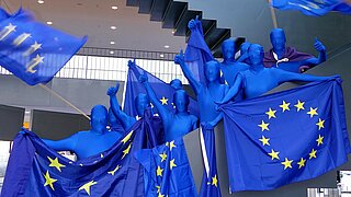 Menschen die einen blauen Ganzkörperanzug tragen und mit EU-Fahnen auf einem Treppenaufgang stehen.