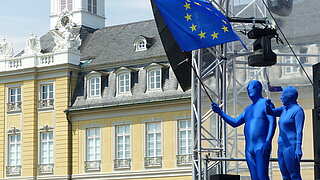 Mann und Frau in blauem Ganzkörperanzug stehen mit Europafahne vor dem Schloss